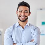 Young happy mixed race businessman standing with his arms crossed working alone in an office at work. One expert proud hispanic male boss smiling while standing in an office